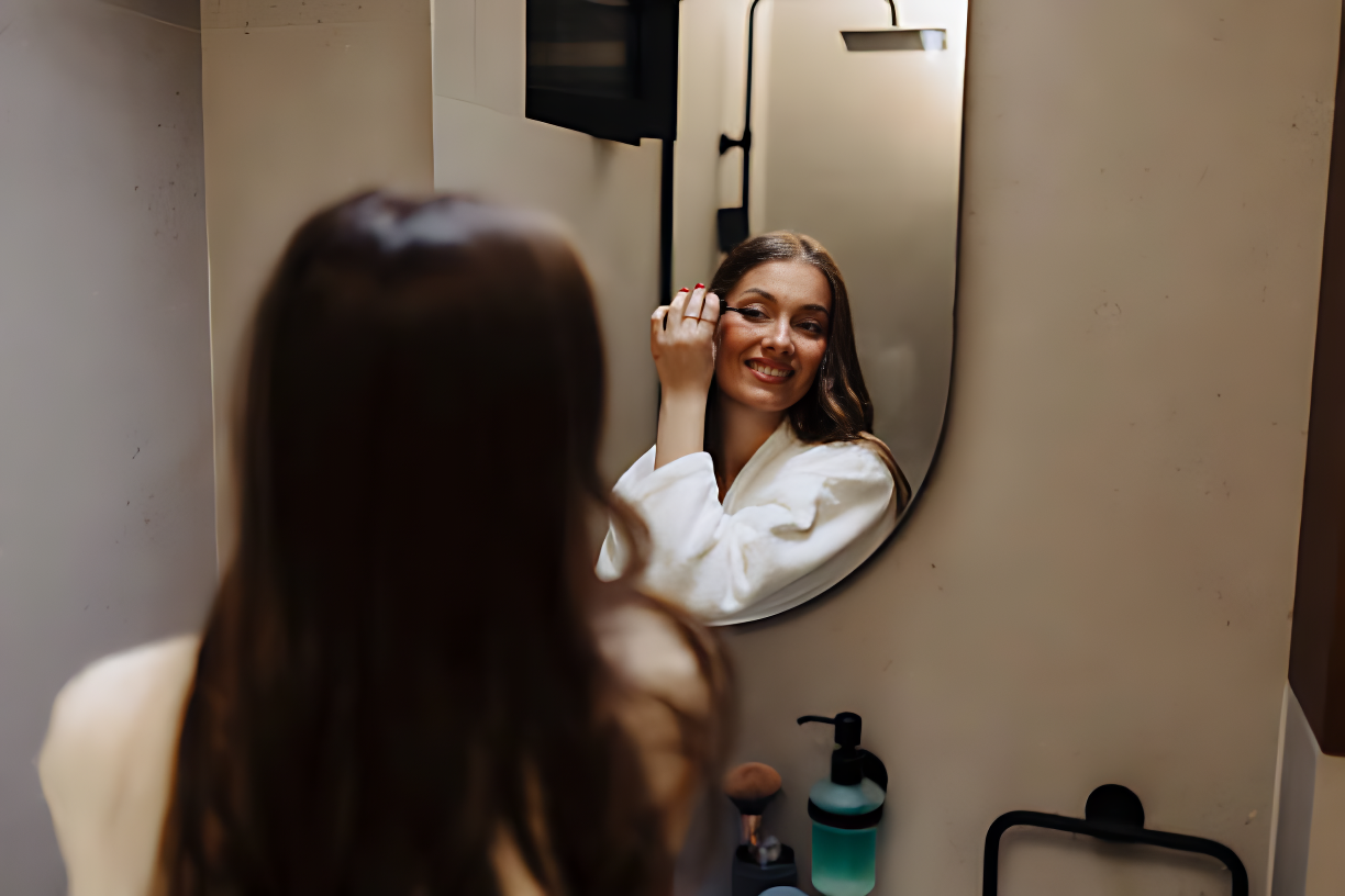 Woman applying makeup in front of a mirror in a bathroom.